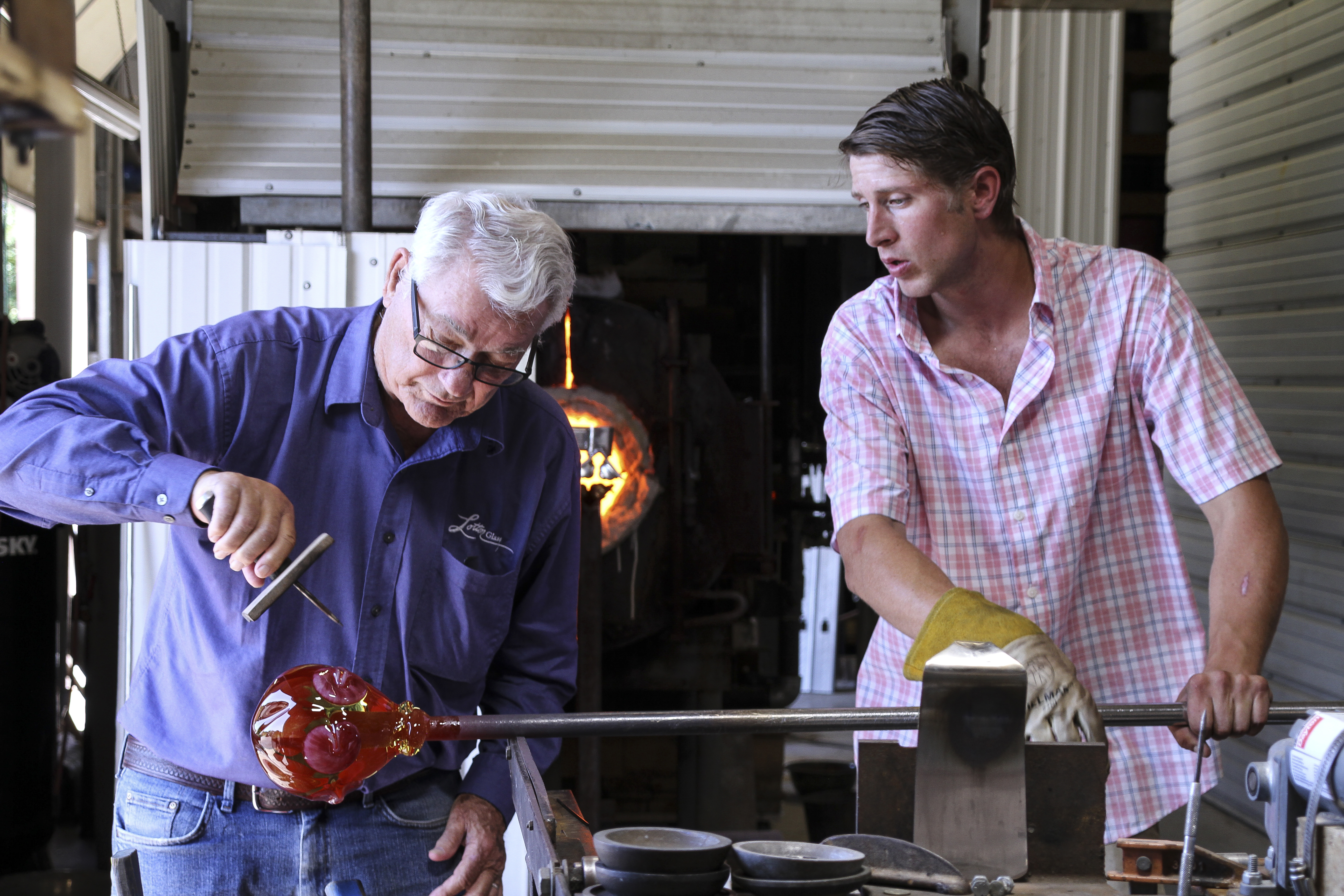 photo of a younger man helping an older man with glass blowing